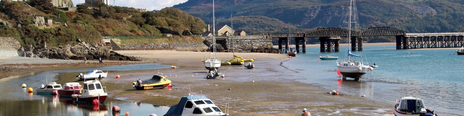 A coastal scene with boats docked at a pier, a sandy beach, and mountains in the background.