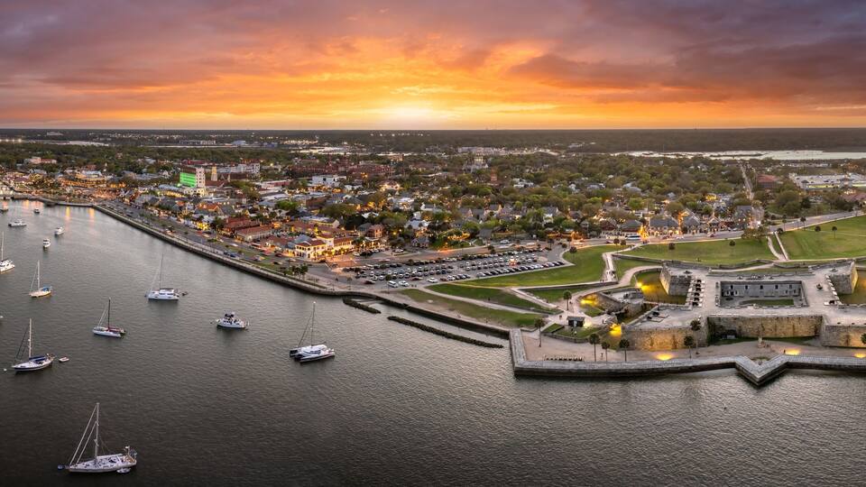 A coastal cityscape at sunset with a prominent fort, marina with sailboats, and a mix of residential and commercial buildings.
