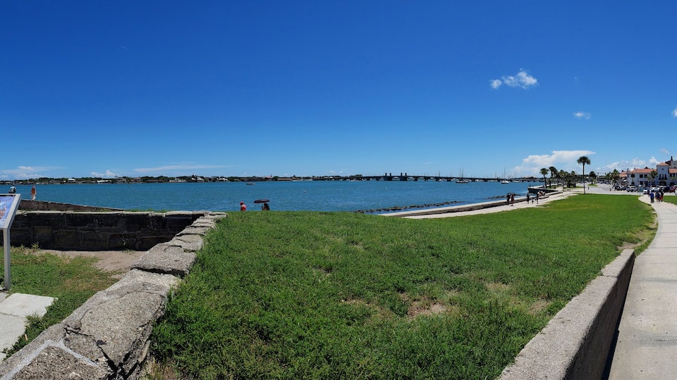 A coastal walkway with a grassy area, a stone wall, and a clear view of the ocean and a bridge in the distance.