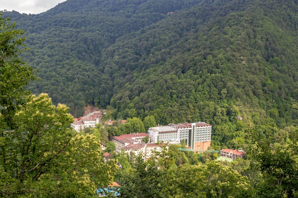 Paesaggio montuoso con fitta foresta e diversi edifici, tra cui un grande albergo e strutture più piccole.