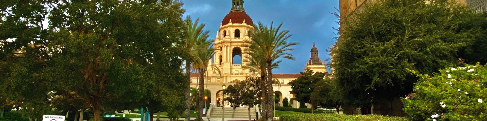 Un edificio histórico con una cúpula y un campanario, rodeado de abundante vegetación y una piscina con una estatua.