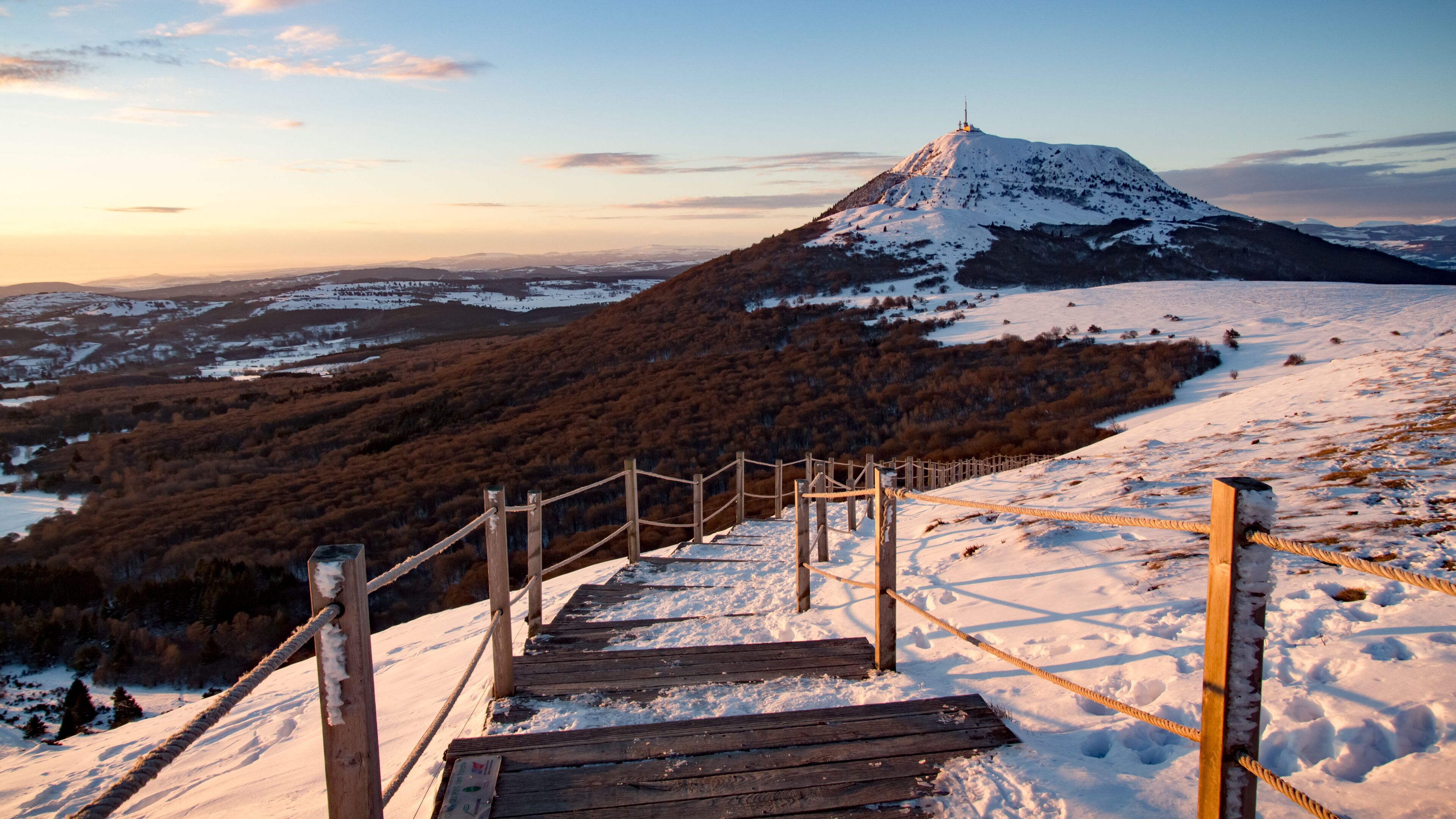 Un escalier en bois menant au sommet d’une montagne enneigée, avec une vallée boisée et un sommet imposant en arrière-plan.