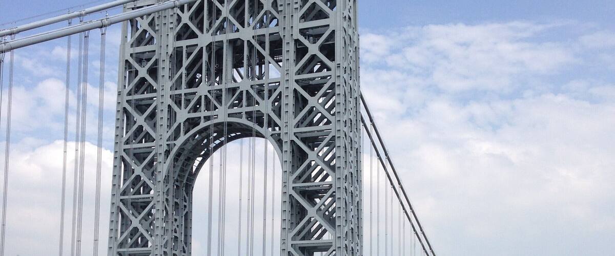 A large suspension bridge with multiple lanes of traffic, a river below, and a cityscape in the background.