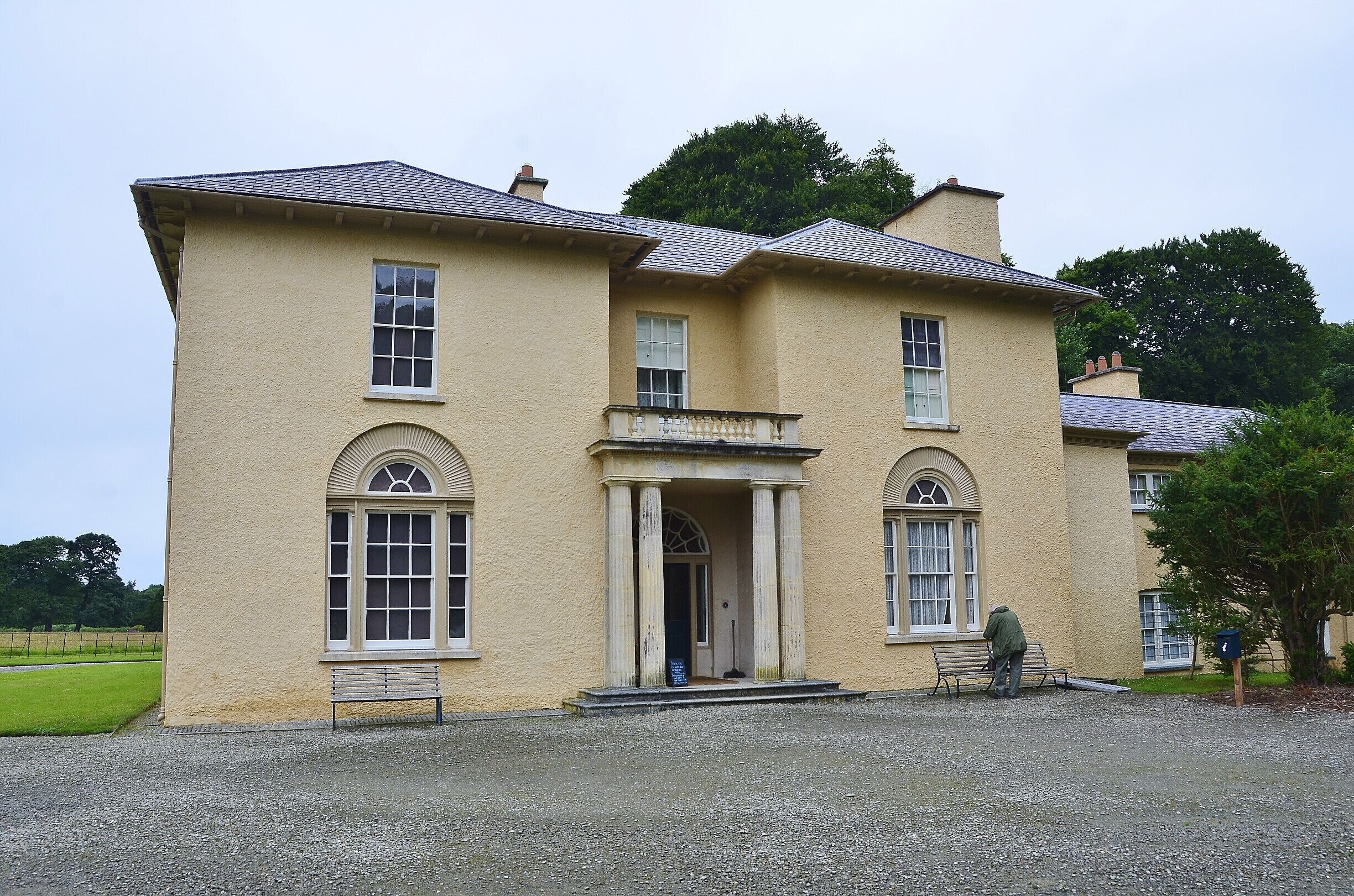 A large, cream-colored building with a symmetrical facade, a central entrance with columns, and a gravel driveway.