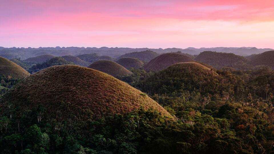 A landscape of rolling hills with dense forests and a small settlement in the foreground.