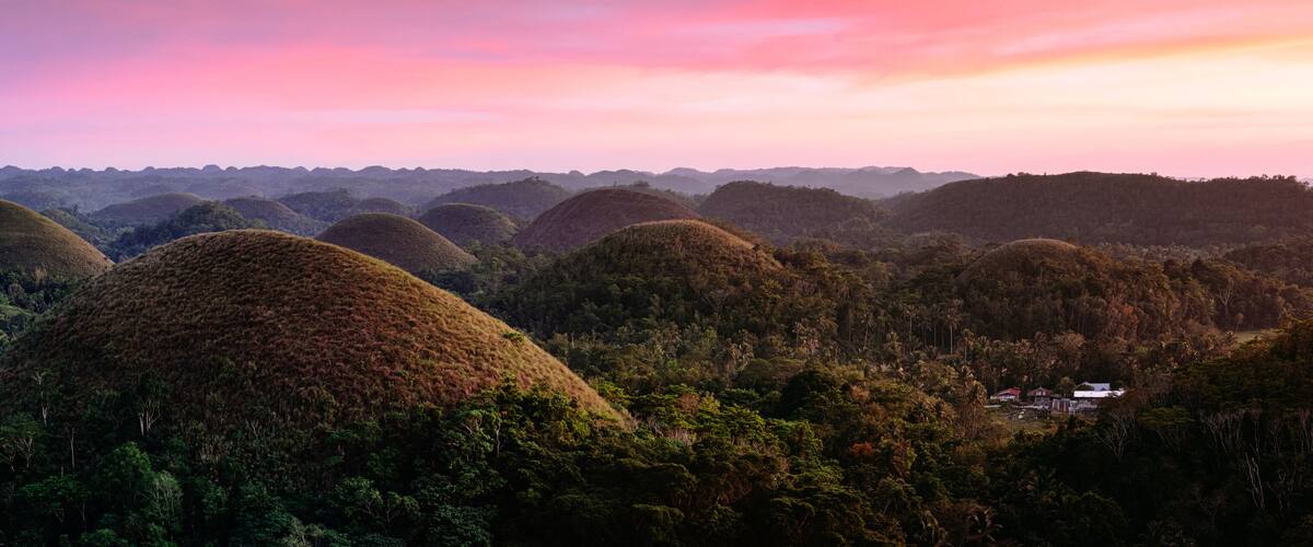 A landscape of rolling hills with dense forests and a small settlement in the foreground.