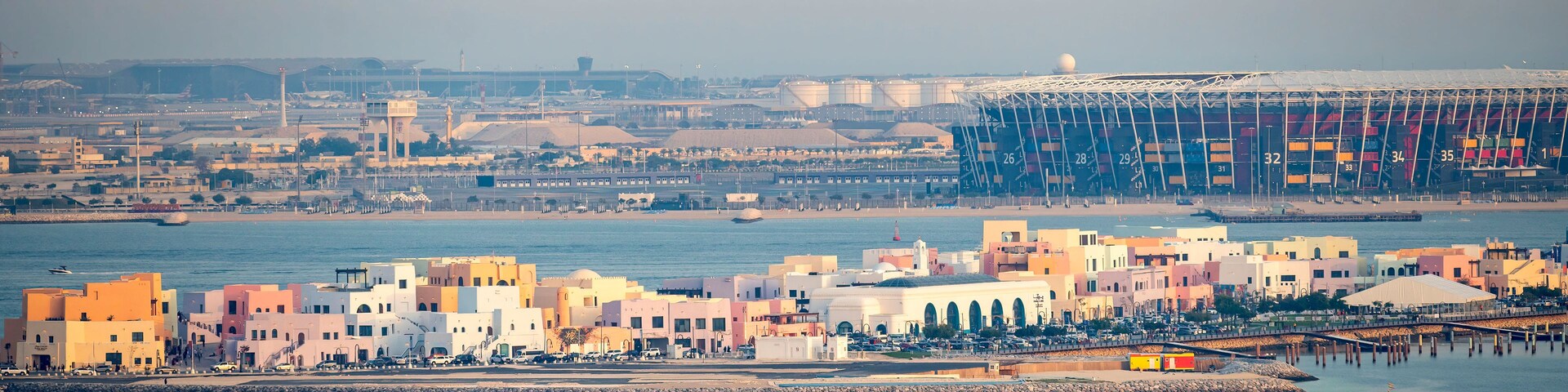 Un paisaje costero con un puerto deportivo, edificios coloridos y una gran estructura industrial al fondo.