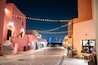 A nighttime street with string lights, buildings with pink and orange facades, and a city skyline in the background.