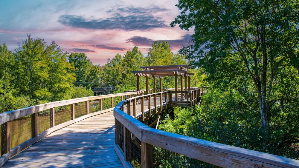 a long winding brown wooden bridge with a pergola over the Sandy Run Creek surrounded by lush green trees and plants with powerful clouds at sunset at The Walk at Sandy Run in Warner Robins Georgia