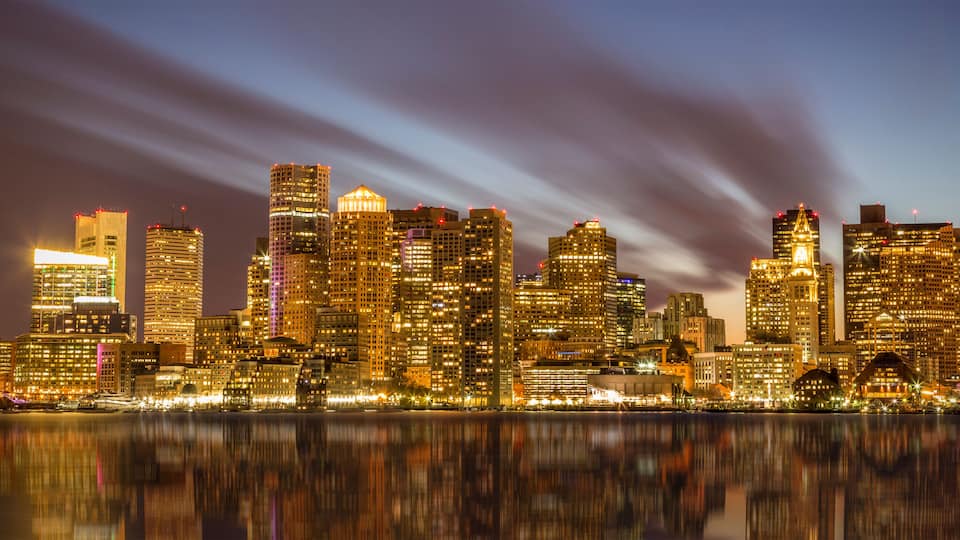 A city skyline at night with illuminated buildings reflecting on the water.