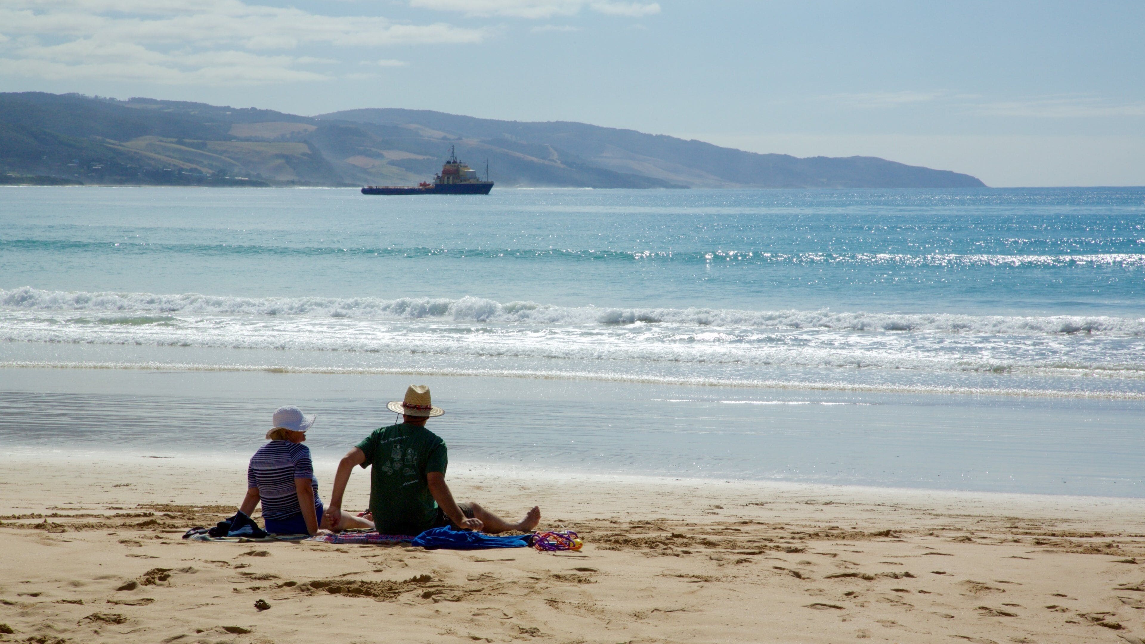 A beach with two people sitting on a blanket, a large ship in the distance, and hills in the background.