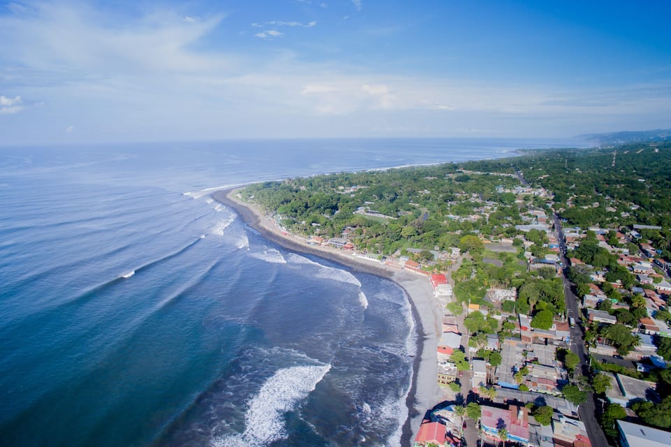 An aerial view of a coastal town with a beach, ocean waves, and dense greenery.