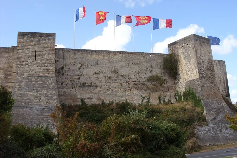 A historic stone wall with flags, including the French and EU flags, and a road in the foreground.