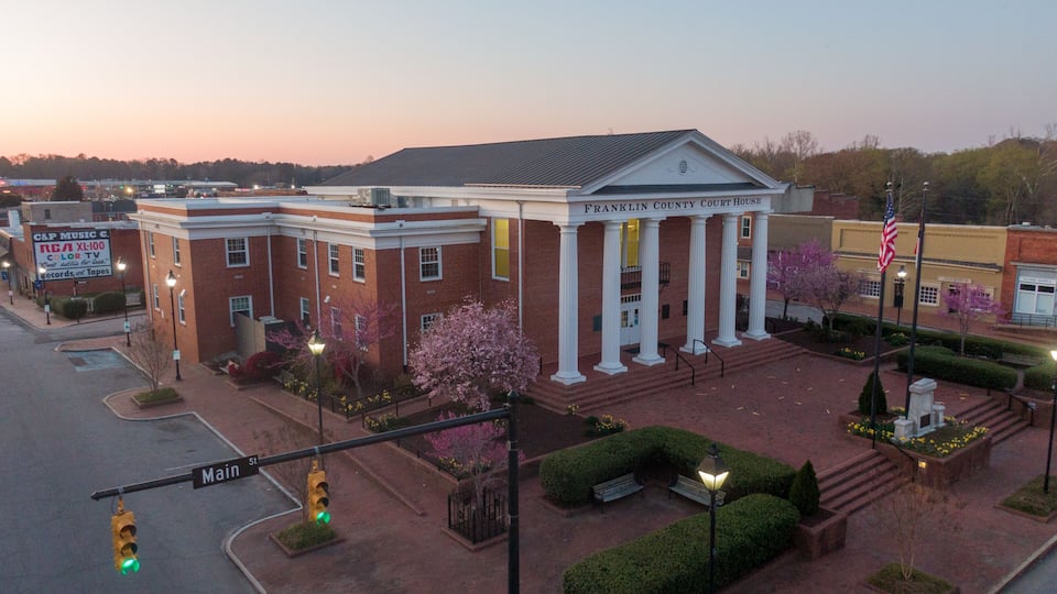 A brick courthouse with white columns and a flagpole, surrounded by well-maintained landscaping and a street intersection.