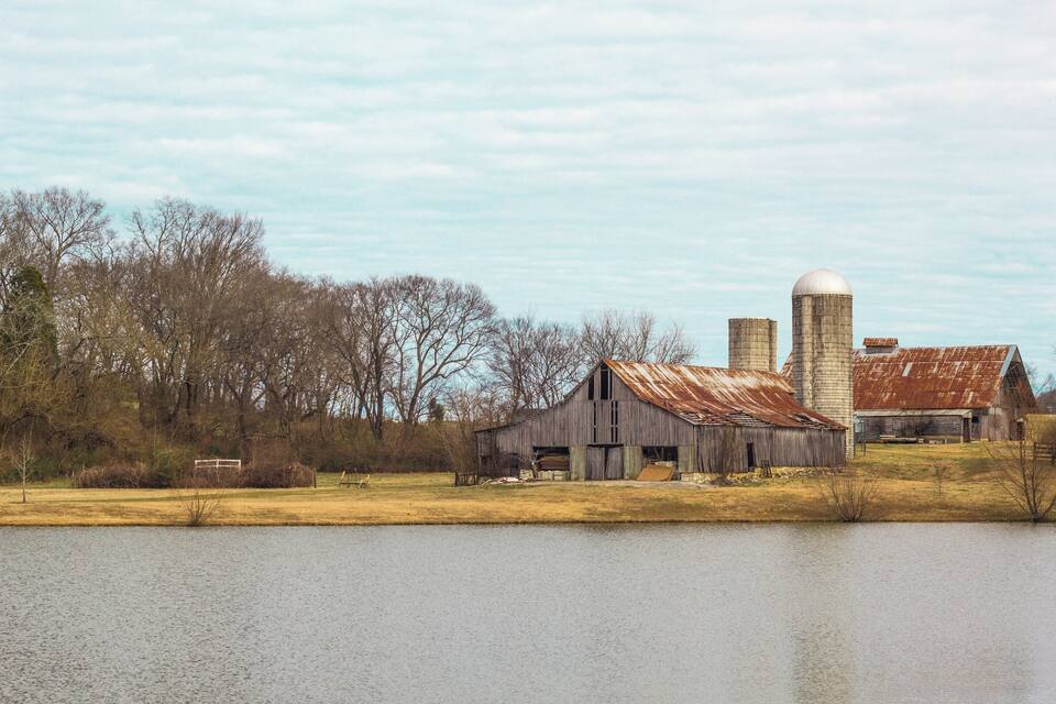 Une grange et un silo rustiques, situés au bord d’un lac paisible, entourés d’arbres sans feuilles et de champs ouverts.