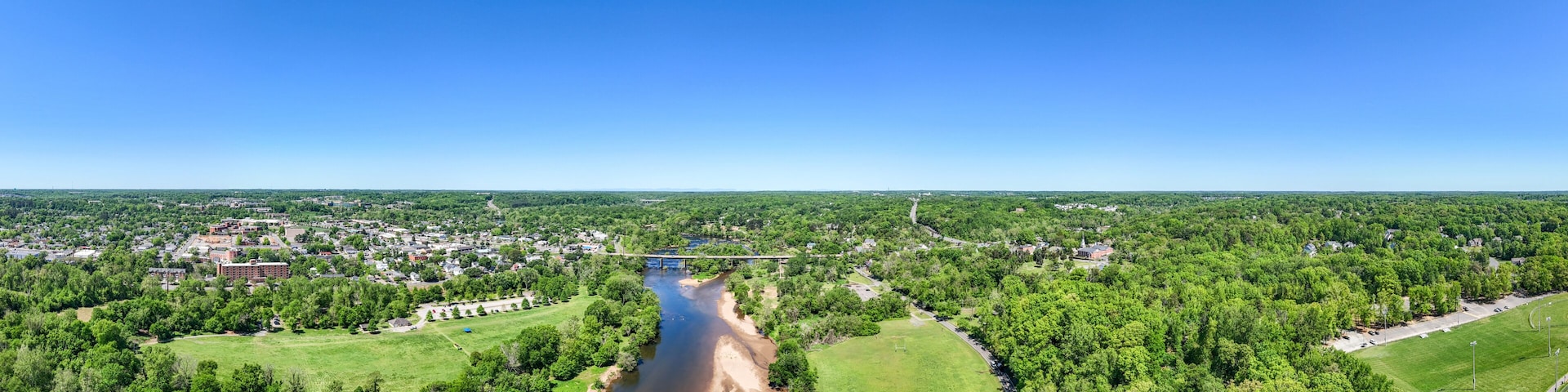A river flowing through a town with a bridge, surrounded by green fields and dense forests.