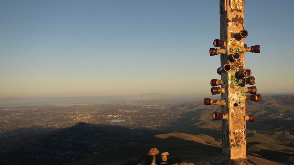 A weathered cross with multiple cups on a mountain peak overlooking a valley.