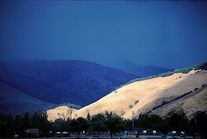 Impending Storm
This picture is from the archives...I think I took this picture in 2002/2003...
En route to running a quick errand in Fremont, CA, where we lived, I was driving by Lake Elizabeth. On the east side of the lake are the Mission Hills. Since I was driving on the west side of said lake, I had an excellent view of the hills, and was witness to an extraordinary sight. Storm clouds coming up from the south had some ridges in complete shadow, while an adjoining ridge, which was not under the influence of the storm yet, was spot lit by sun. I could see the storm traveling north rapidly, and I knew that the situation would not last. Parking the car, I raided my camera bag for my camera and a telephoto, and with tripod in hand, ran to the water's edge. I managed to setup and expose a few frames, before the sunlit ridge came under the shadow of the storm. The magic was gone and the entire landscape turned a uniform dull gray. 
I don't remember any technical details other than that the camera used was a Nikon F100. I owned the Nikkor 80-200 mm f2.8 lens then, which I was partial to, so that may well have been my lens of choice for this shot. Similarly, I used to shoot with Fuji transparencies at the time, so my guess is that I used Fuji Velvia for this shot.