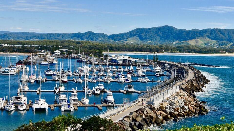 A marina with numerous boats docked, surrounded by rocky breakwaters and a backdrop of mountains and a beach.