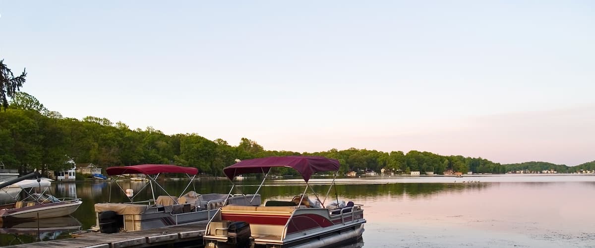 A calm lake with boats docked at a wooden pier, surrounded by trees and a distant shoreline.