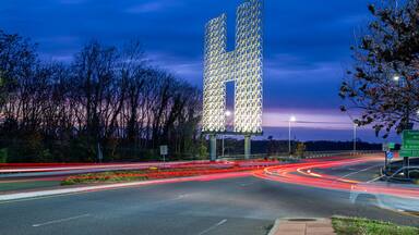 Um entroncamento rodoviário com uma estrutura iluminada peculiar em forma de 'U', rodeado por árvores e placas de sinalização.