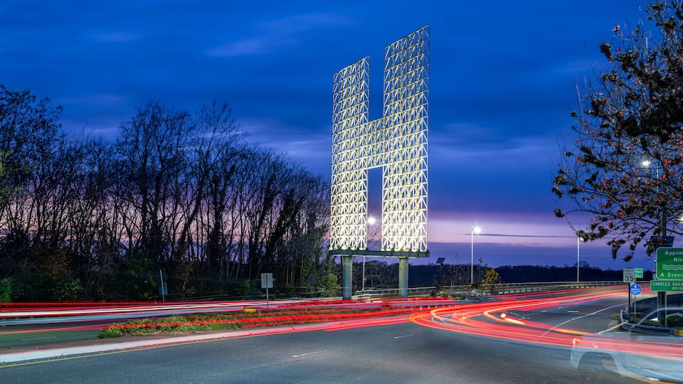 A highway interchange with a distinctive illuminated structure resembling a 'U' shape, surrounded by trees and featuring road signs.