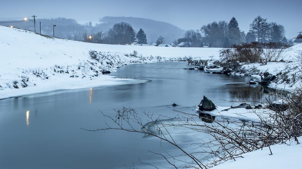 A snow-covered river with a bend, surrounded by trees and a distant mountain range.