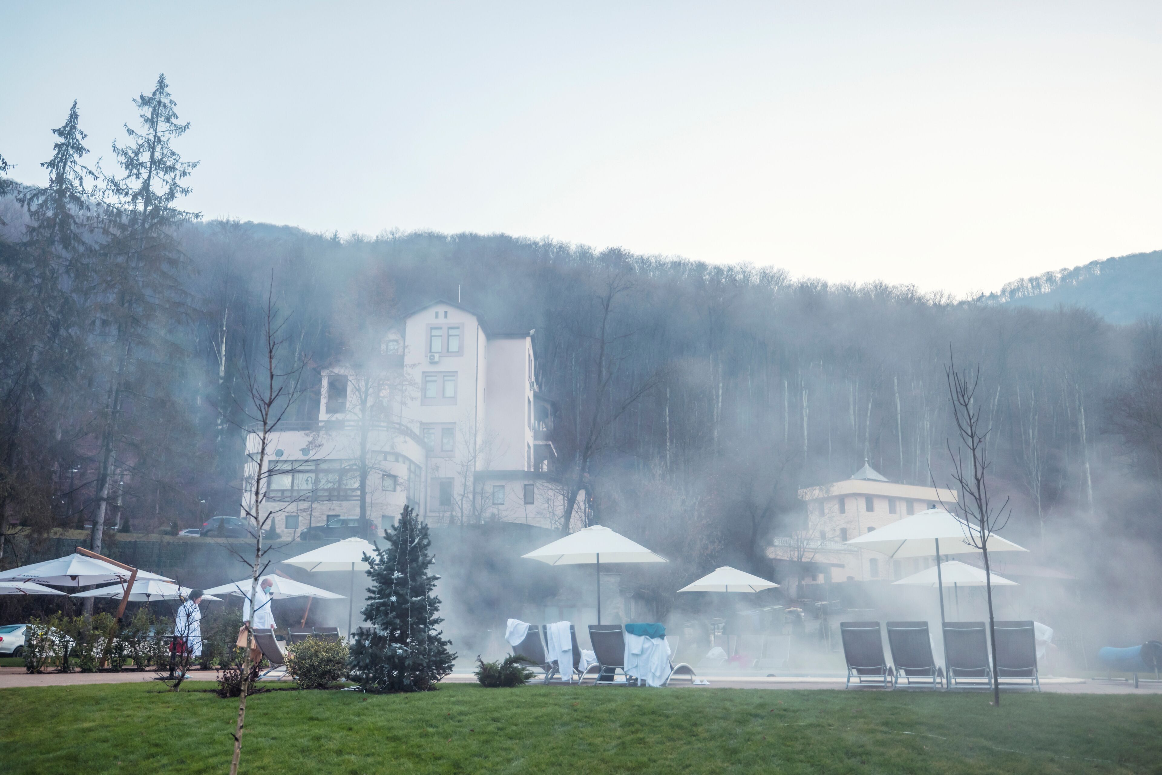 Steam rises gently from the hot springs, enveloping cozy lounging areas and elegant buildings, while the serene mountains stand guard against the early morning sky