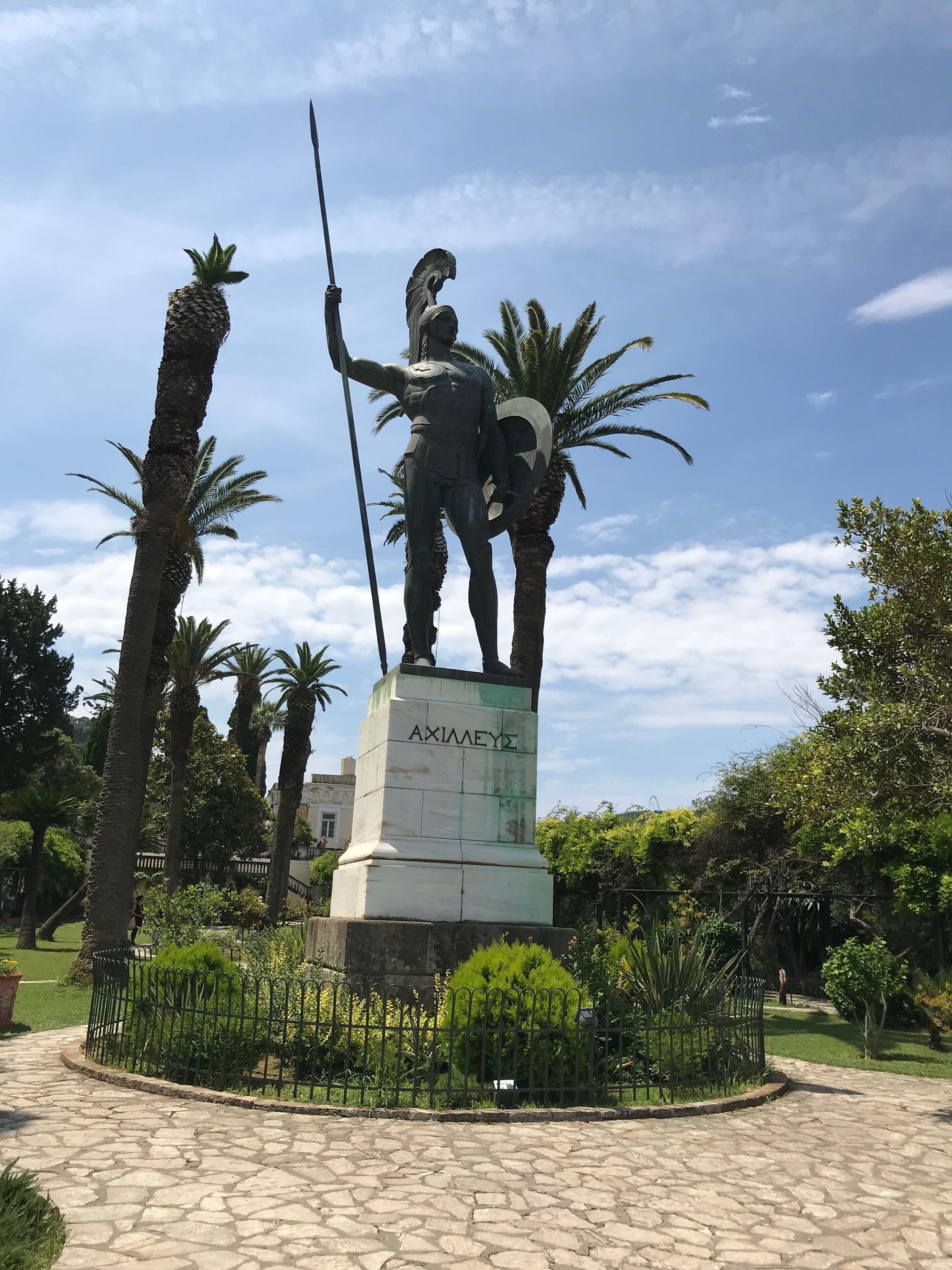 Una estatua de un guerrero sosteniendo una lanza y un escudo, con la inscripción "AXIMETEY", rodeada de palmeras y un camino empedrado.