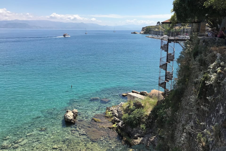 A coastal landscape with clear turquoise waters, rocky shorelines, and a distant sailboat.