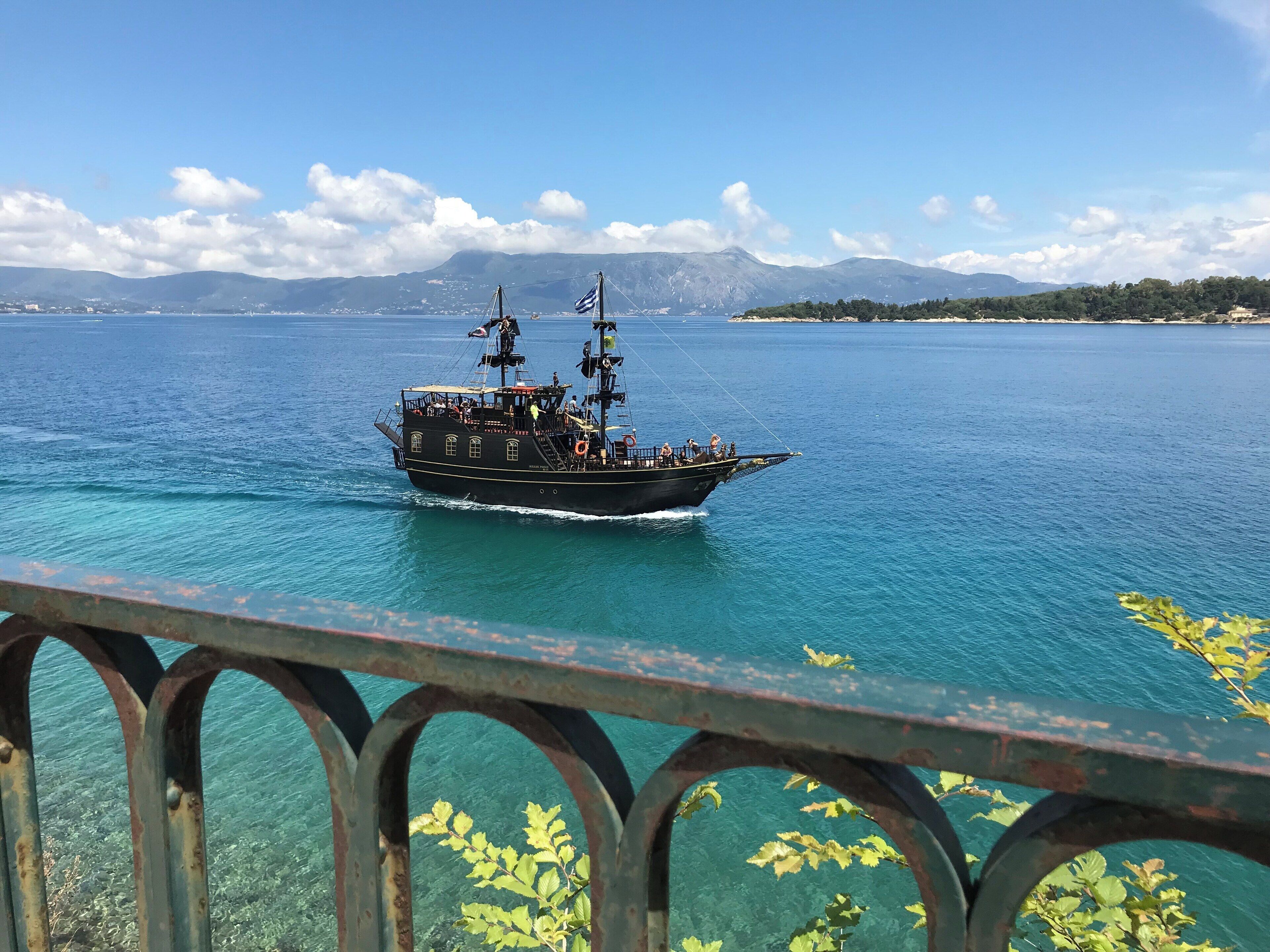 A black ship with people on deck, sailing on a clear blue sea with mountains in the background.