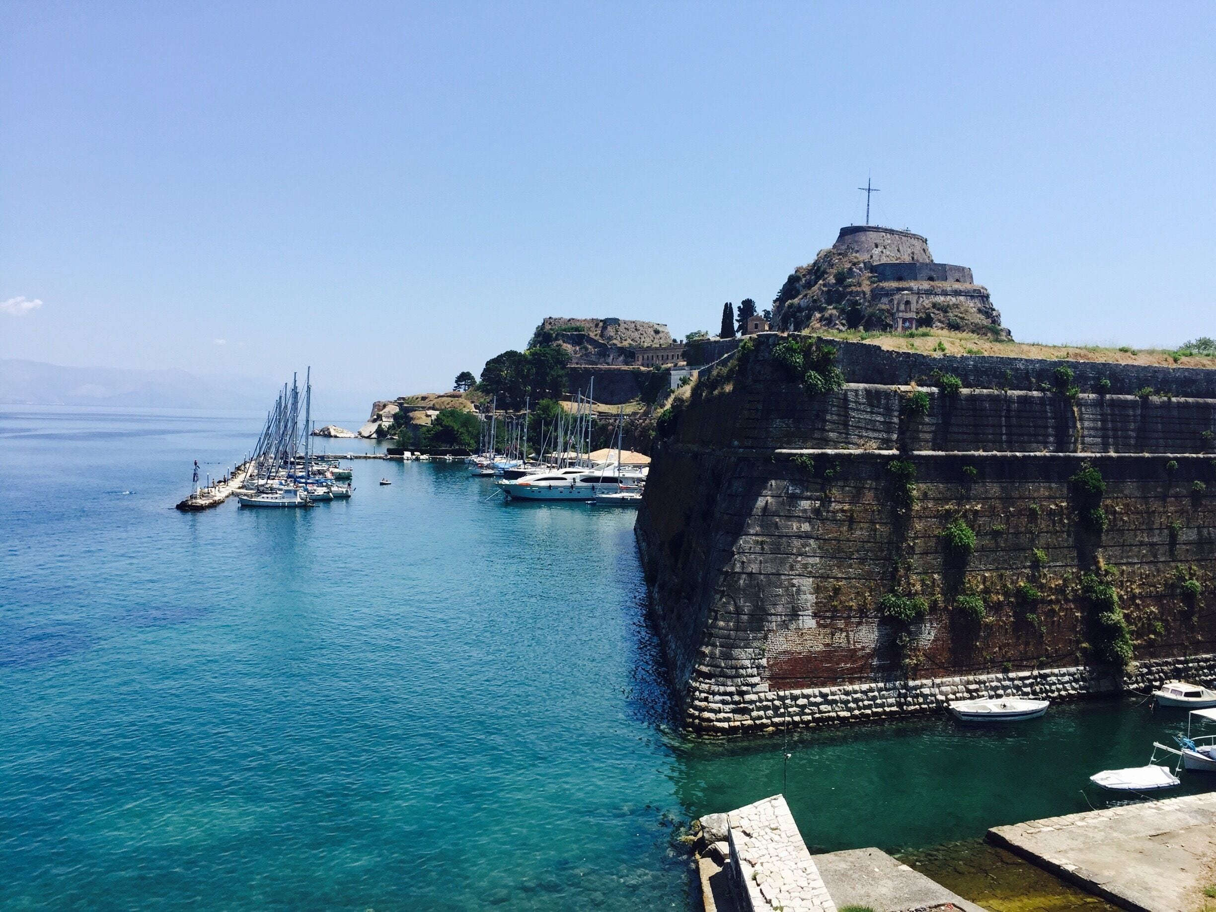 A coastal scene with a fortified structure, a clear blue sea, and several boats.
