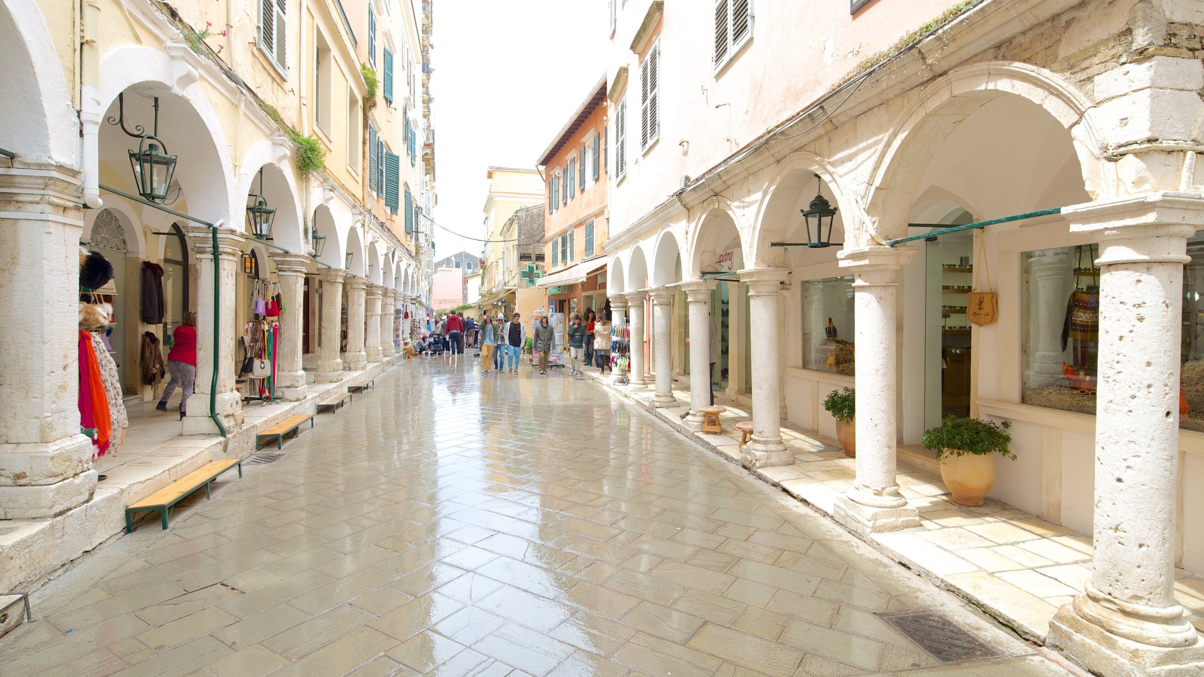 Uma rua de paralelepípedos molhada, ladeada por edifícios brancos, arcos e lojas.
