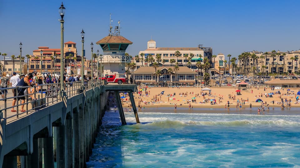 A pier extending into the ocean with people walking along it, a beach with palm trees and umbrellas, and buildings in the background.