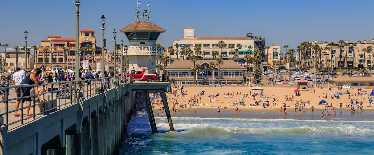 A pier extending into the ocean with people walking along it, a beach with palm trees and umbrellas, and buildings in the background.