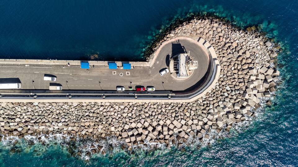 An aerial view of a coastal road with a roundabout, parked cars, and a rocky breakwater.