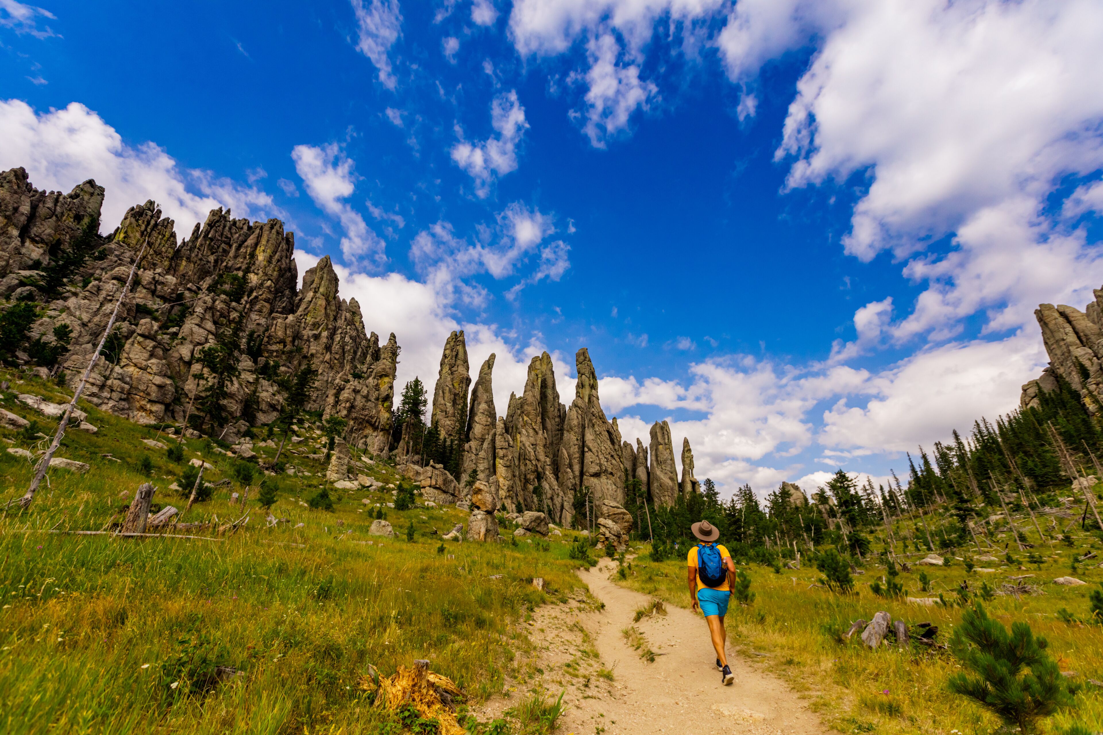 A hiker on a dirt path surrounded by tall, rugged rock formations and dense forest under a partly cloudy sky.