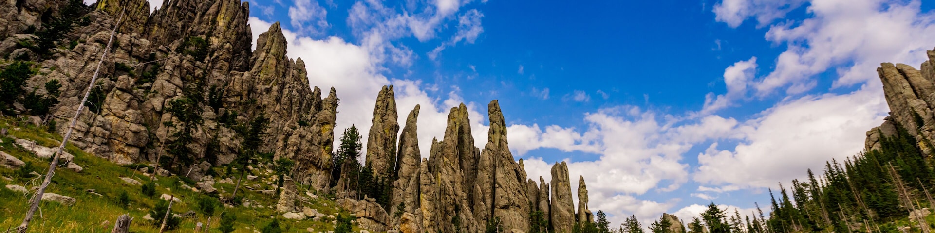 Un excursionista en un sendero de tierra, rodeado de altas y escarpadas formaciones rocosas y un bosque denso bajo un cielo parcialmente nublado.