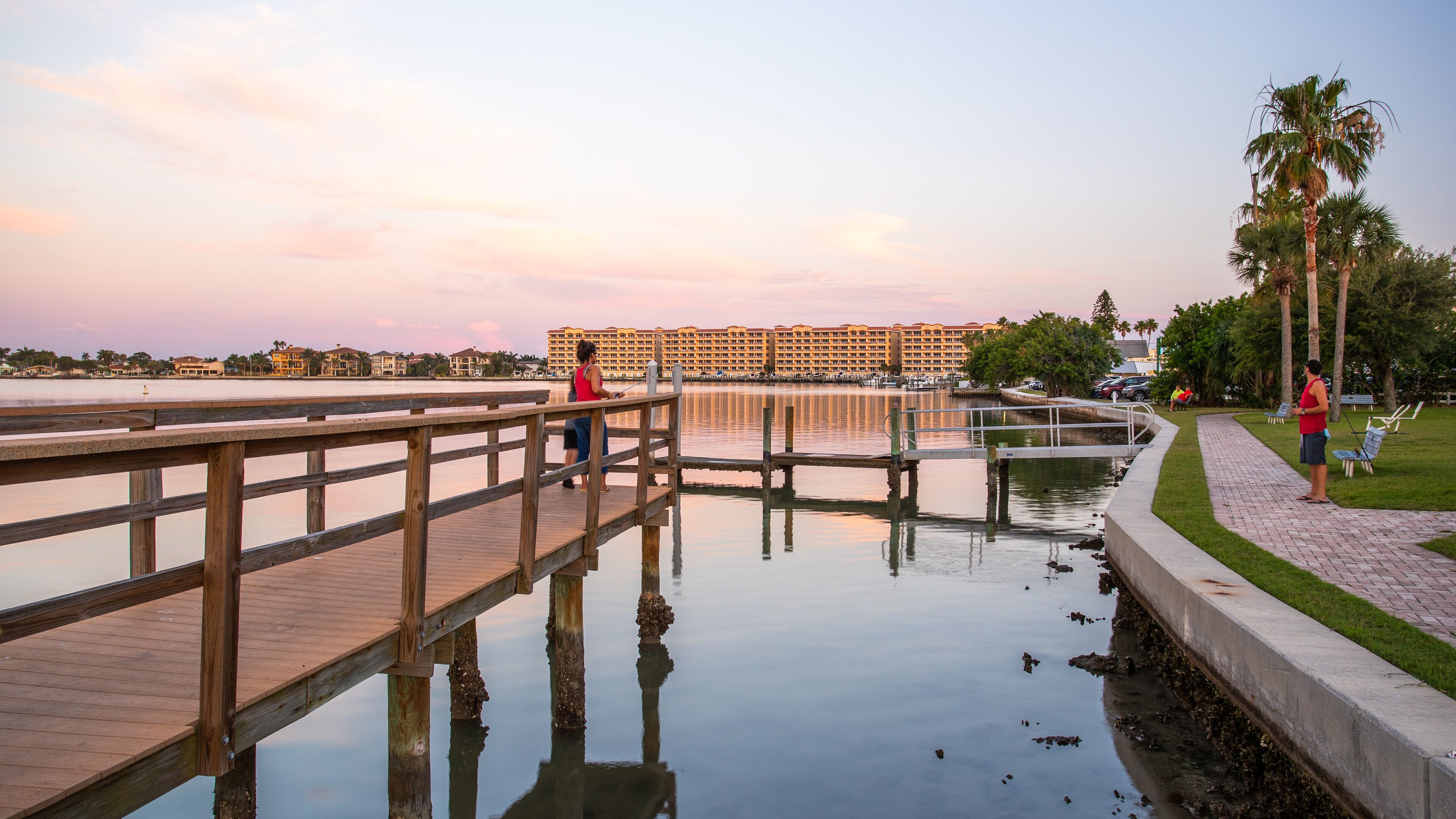 A wooden boardwalk over calm water with reflections, palm trees, and a hotel in the background.