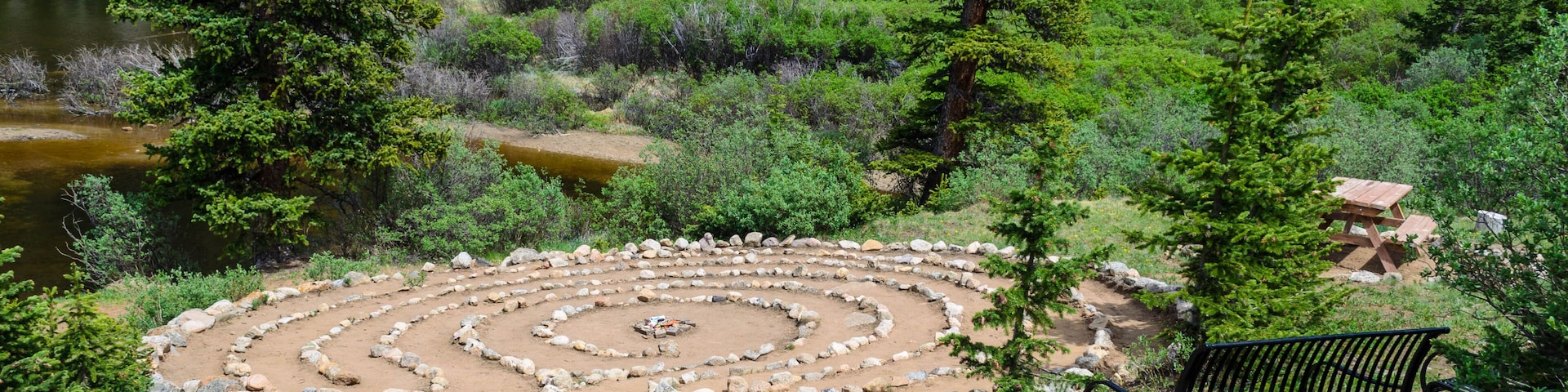 A circular stone labyrinth, a bench, and a forested area with a lake.