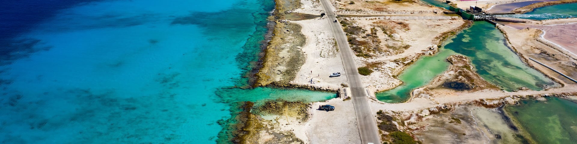 An aerial view of a coastal area with pink and purple salt flats, clear blue waters, and a road running parallel to the shore.