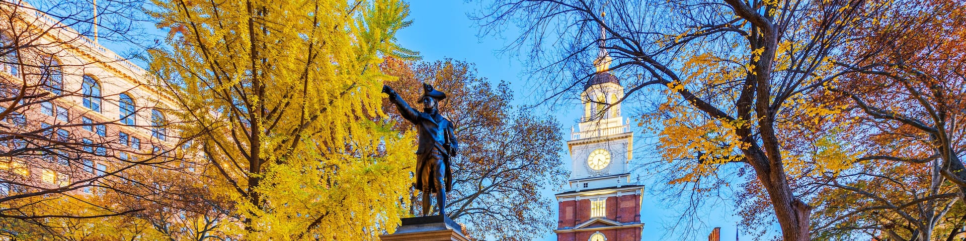 A statue in a park with autumn trees and a historic building with a clock tower.
