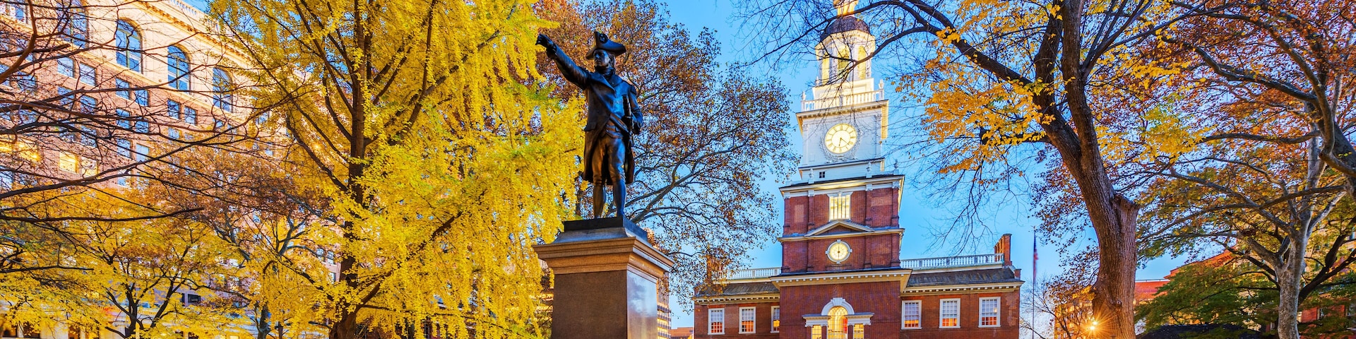 Uma estátua em um parque com árvores de outono e um edifício histórico com torre do relógio.