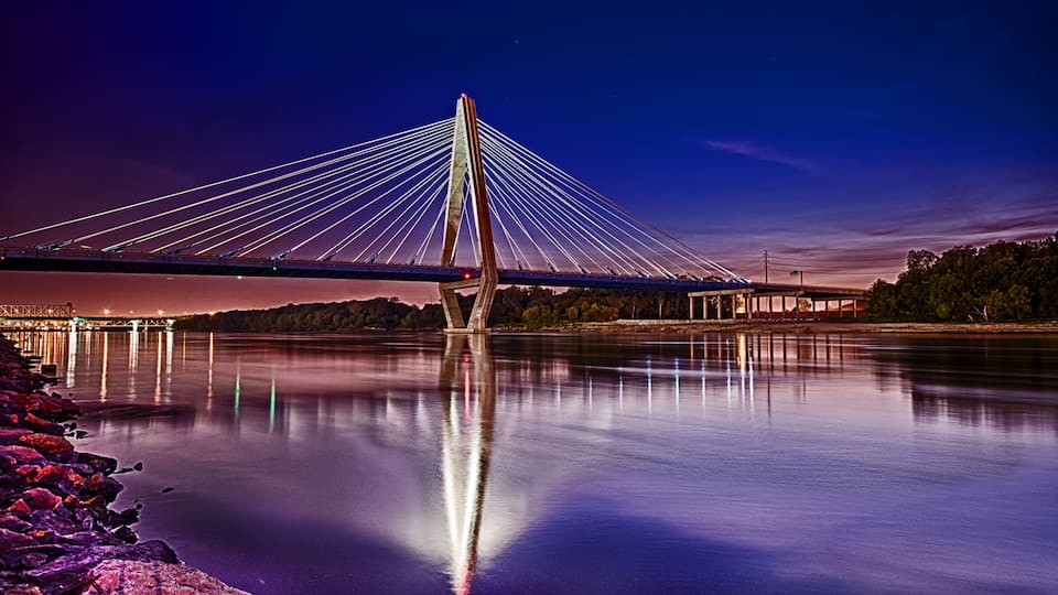 A cable-stayed bridge at dusk with its cables illuminated, reflecting on the water.