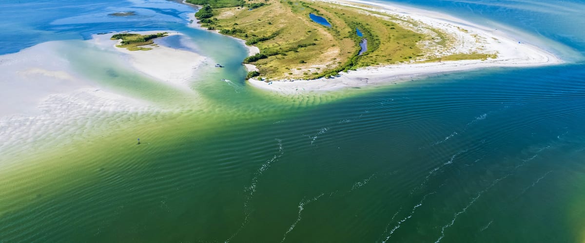 An aerial view of a coastal landscape with a sandy beach, a small island with vegetation, and clear blue waters.