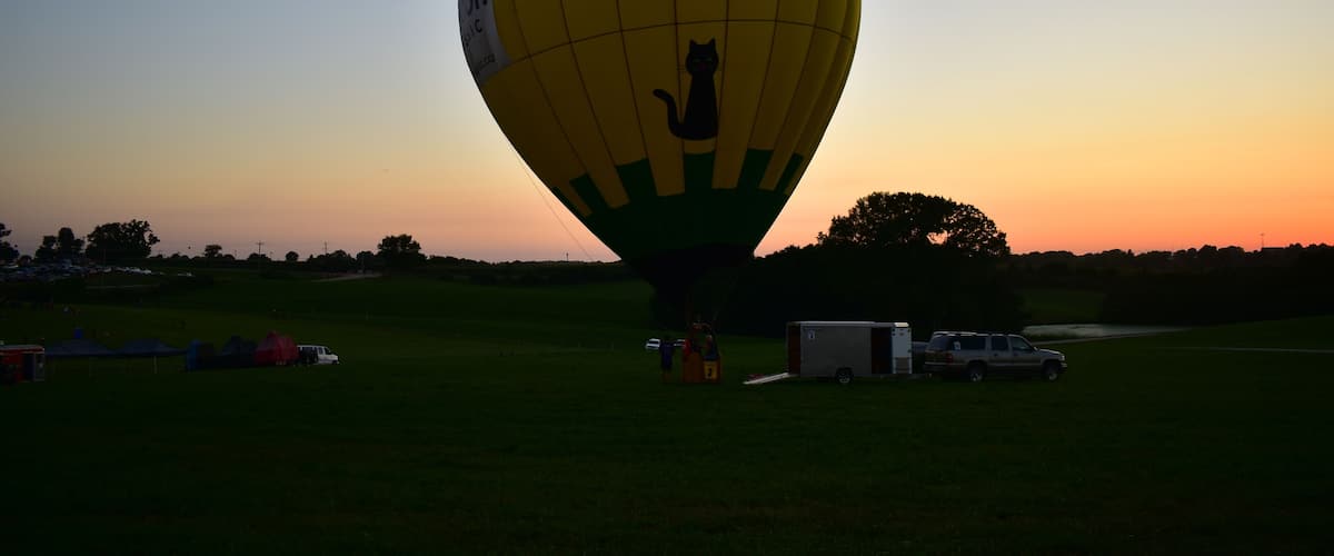 A colorful hot air balloon with a cat silhouette, parked in a grassy field with tents and vehicles in the background.