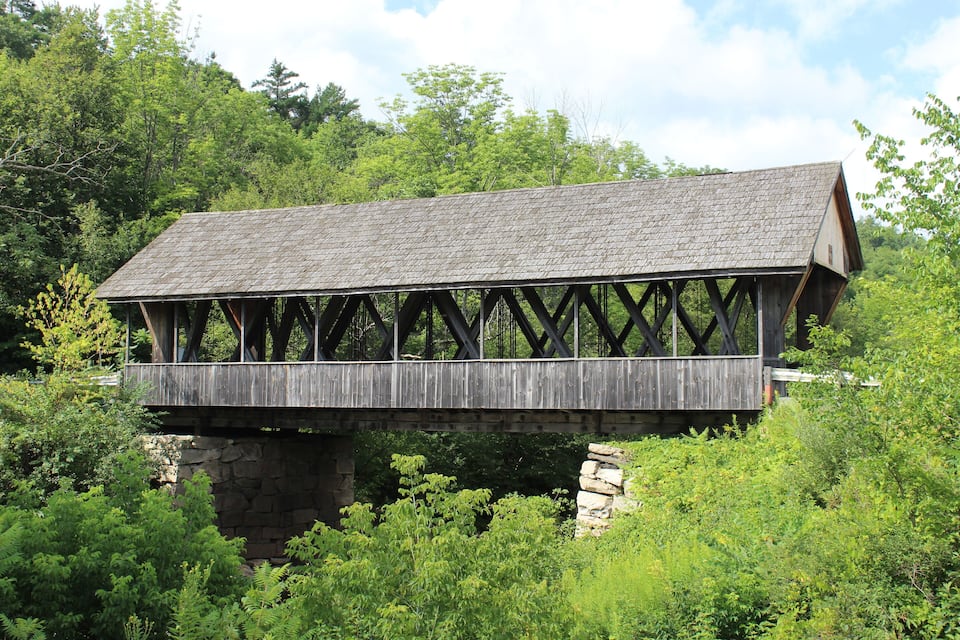 A wooden covered bridge with a stone foundation, surrounded by lush greenery and trees.