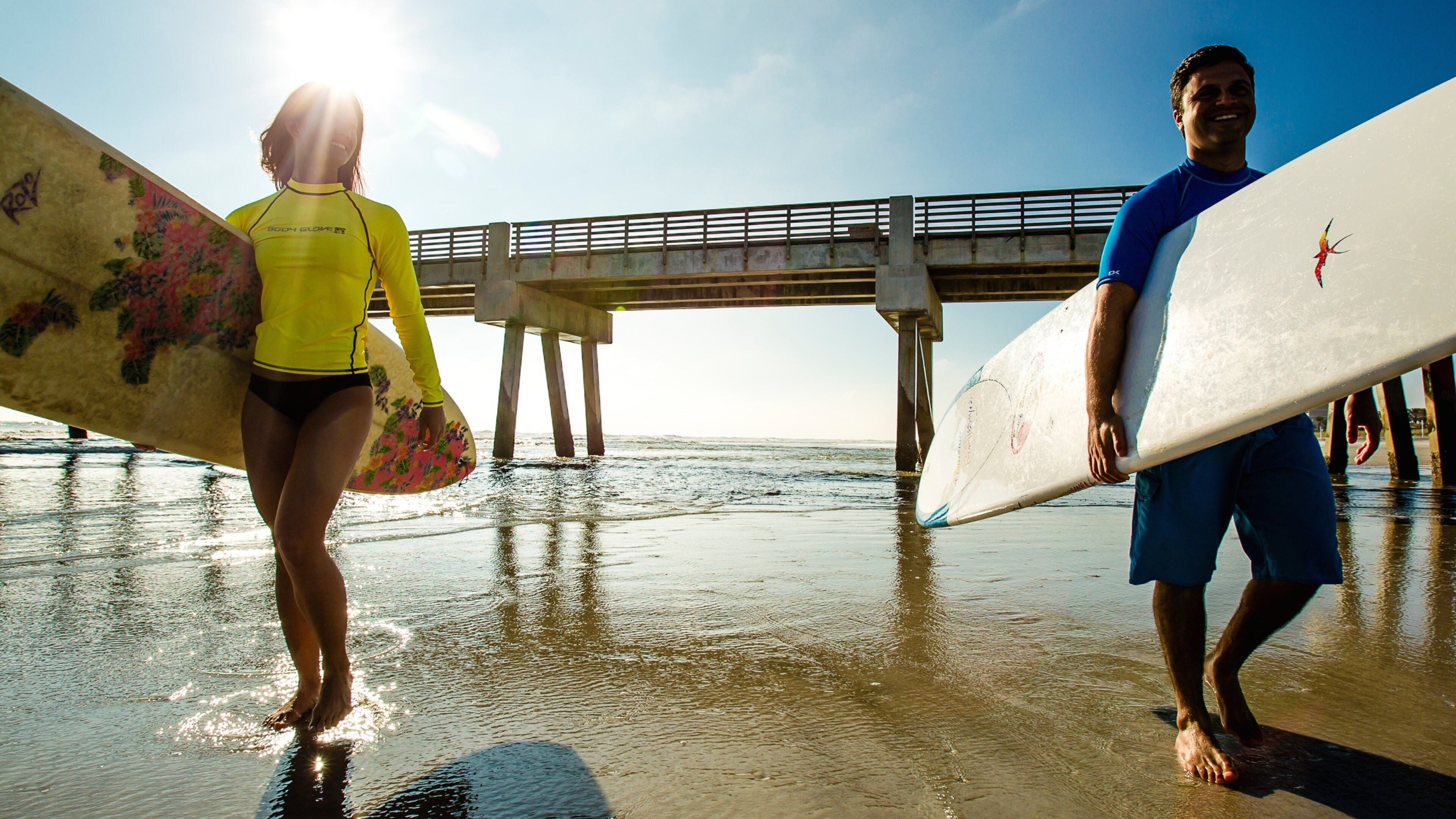 Two people carrying surfboards under a bridge at the beach.