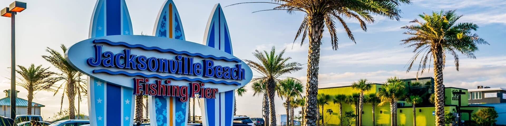 A sign for Jacksonville Beach Fishing Pier with palm trees and parked cars.