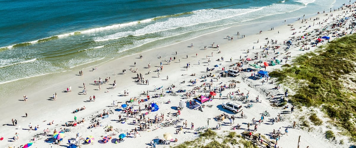 A crowded beach with people, umbrellas, and a pier in the background.